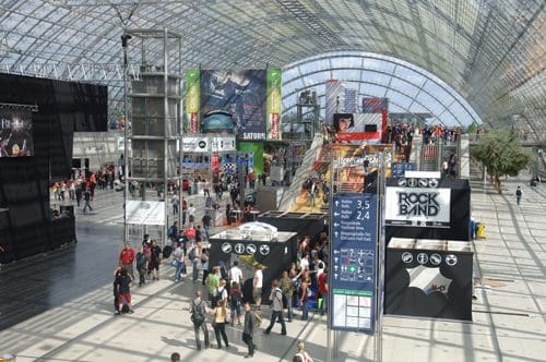 A bustling exhibition center filled with visitors, showcasing various booths and displays under a large glass roof. People navigate the space, with signs for events like 'Rock Band' visible.