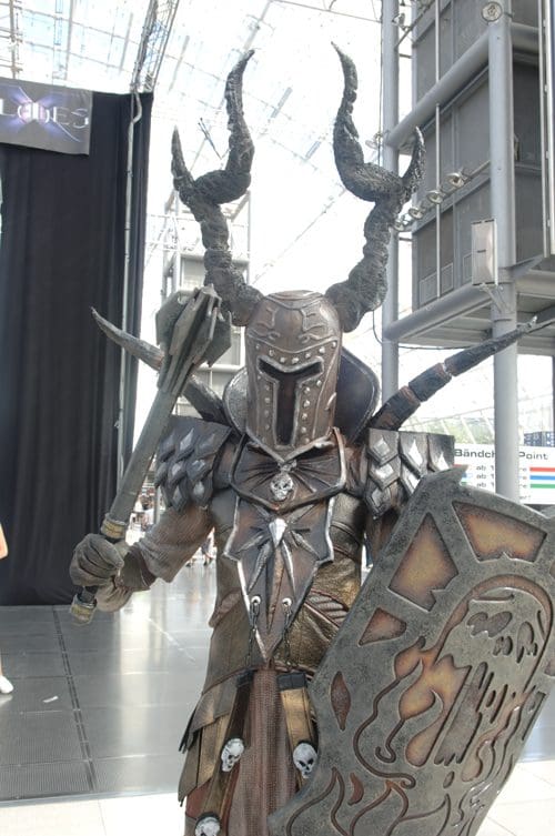 A cosplayer dressed in a detailed, dark fantasy armor with horns, holding a spear and a shield, poses confidently at a convention center. The background features glass walls, suggesting a modern venue.