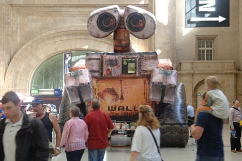 A large cardboard cutout of the animated robot WALL-E stands in a busy train station, surrounded by people walking by. The robot features distinctive large eyes and a weathered, rusty appearance, creating a whimsical focal point.