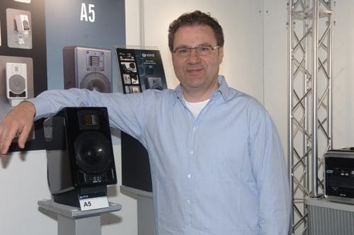 A man in a light blue shirt stands next to a black studio monitor speaker labeled 'A5' displayed on a stand. Behind him, there are images of other speaker models on a white wall. The setting appears to be an exhibition space.