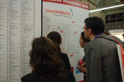 A group of four people examining a large information board displaying a map and listings at an event venue. The board includes labels like 'Thomson' and 'Information Point', indicating directions and services.