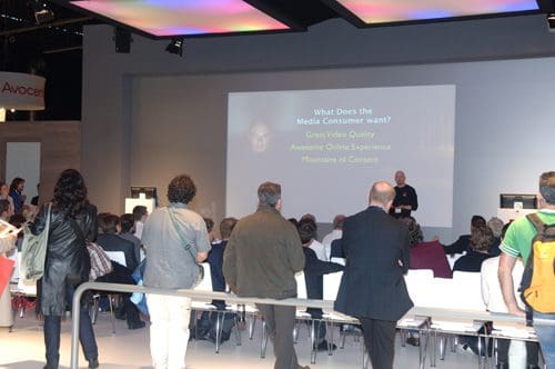 A crowd of attendees gathered in a conference hall facing a speaker presenting on stage. The presentation slide shows the topic 'What Does the Media Consumer Want?' with key points about video quality and online experiences.