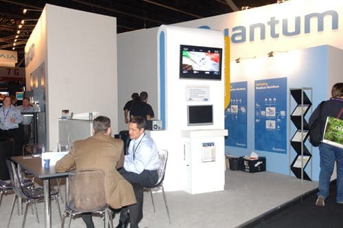 A modern exhibition booth labeled 'Quantum' showcasing a sleek presentation area. Two men are seated at a table engaging in conversation, while a television displays visuals on the booth's wall. Additional informational displays are visible.