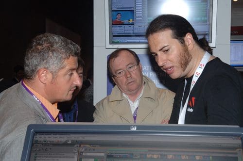 Three men engaged in a discussion at an exhibition booth, with one man gesturing as he explains something. A computer screen is visible in the foreground showing software interface.
