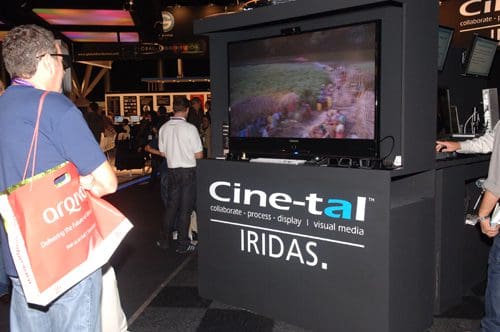 A man observing a large display showcasing colorful visuals at a trade show booth with the name "Cine-tal IRIDAS" prominently featured. Attendees are gathered around in a busy exhibition space.