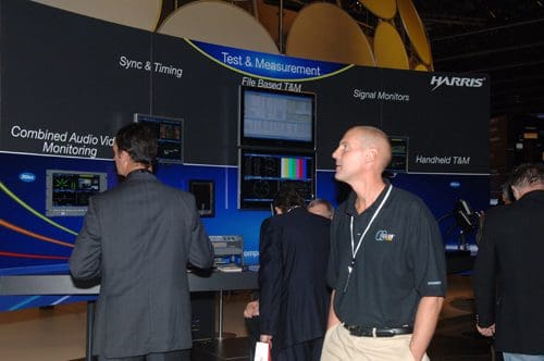 Several professionals observing technology displays at a trade show booth featuring information on test and measurement equipment, including screens for signal monitoring and audio video monitoring.