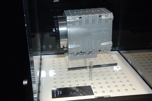 A close-up of a silver automotive supercharger displayed in a showcase, with visible ventilation slots and a polished surface, resting on a lighted platform. A product card is positioned beside it.