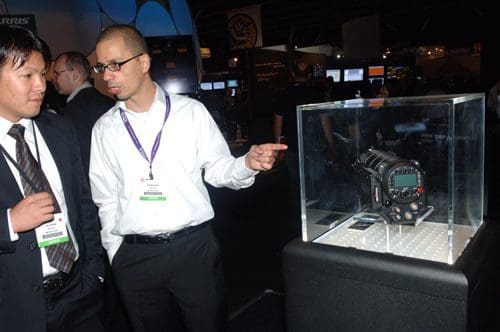 Two men in business attire engaged in conversation, standing near a transparent display case. Inside the case, a high-tech camera with various buttons and a digital display is prominently featured on a light base.