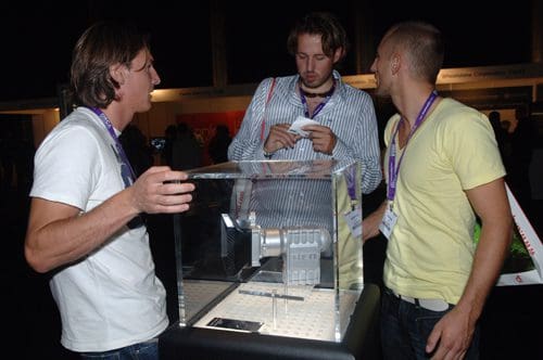 Three men engaged in conversation while examining a clear display case containing a model or exhibit at an event. They appear focused and curious, with event badges visible around their necks.