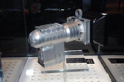 A close-up view of a metallic silver turbocharger displayed in a glass case, with intricate detailing on its surface. The turbocharger features a cylindrical shape with an attached rectangular component, showcasing its engineering design.