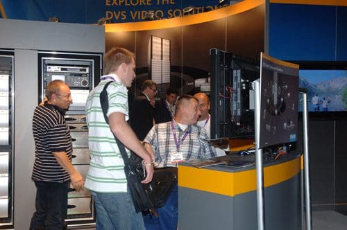 A group of attendees at a technology exhibition interact with a display featuring video solutions. One person examines a monitor while others engage in discussions near equipment racks.
