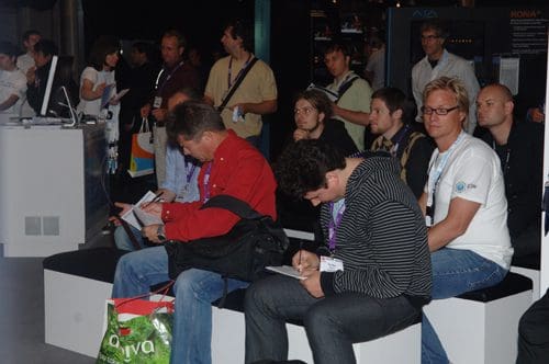 A group of people seated in a technology event venue, engaged in discussions and taking notes. The audience consists of men and women, with some wearing badges and casual attire. A digital presentation is visible in the background.