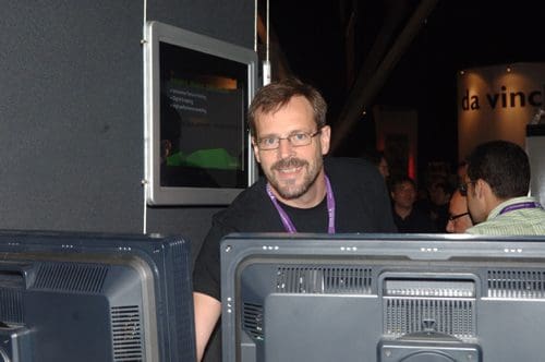 A smiling man with glasses, wearing a black shirt, stands behind two computer monitors at a tech event. In the background, several people engage with presentations or displays.