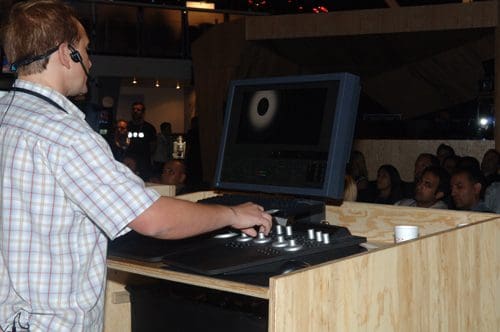 A man operating a control panel with buttons and dials, focused on a large monitor displaying a circular graphic. In the background, an audience watches intently in a well-lit venue.