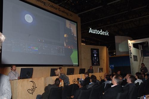 A digital presentation taking place at a technology event, featuring a large screen displaying a moon image and various editing tools. An audience sits attentively in front of a panel, while branded booths are visible in the background.
