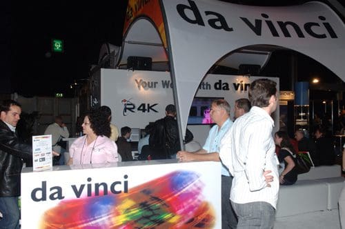 A trade show booth for 'da vinci' featuring a colorful logo. Several attendees engage with staff at the reception, while a large screen displays digital content in the background.