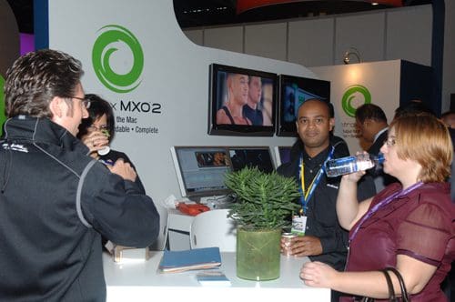 A group of professionals engaged in conversation at a tradeshow booth. One person drinks water while others discuss nearby displays, with a potted plant on the counter and video screens showing content in the background.