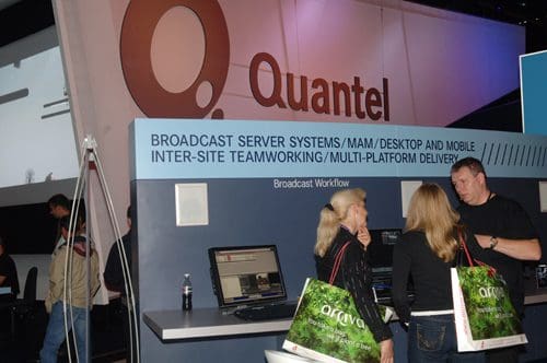 A trade show booth for Quantel featuring a large logo and a display about broadcast server systems. Two individuals are engaging in conversation, while another person examines a computer demo on the counter.