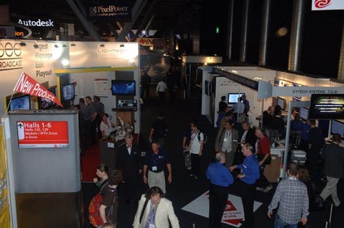 A busy exhibition hall with various booths featuring technology displays. Attendees are walking between the booths, engaging in conversations. Signs overhead indicate different areas of the event.