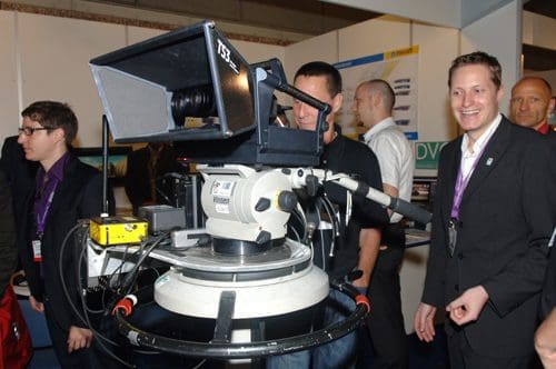 A television camera on a rotating tripod, with an operator behind it, set up at an exhibition. Two men in suits smile in the foreground, while others engage in discussions in the background, surrounded by display stands.