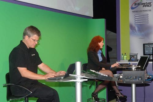A man and a woman working at computers on a stage with a green backdrop. The man, wearing glasses and a black shirt, focuses on his laptop, while the woman, dressed in a black suit, operates a desktop setup. Bottled water is visible on the table.