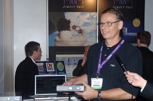 A man holding a device at a trade show booth with a smile, wearing glasses and a name tag. Behind him are promotional materials and a laptop. Others are engaged in conversation in the background.