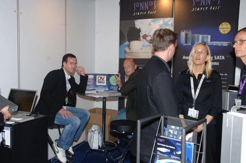 A trade show booth featuring several attendees engaged in conversation. Two men sit at a table, one looking thoughtfully, while a woman speaks to a group. The backdrop displays promotional banners and products.