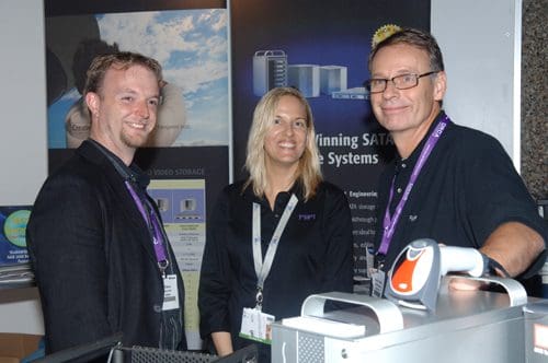 Three professionals posing at a trade show booth. The man on the left has short hair and is wearing a black jacket, while the woman in the middle has long blonde hair and wears a black shirt. The man on the right wears glasses and a dark shirt, with promotional materials displayed behind them.
