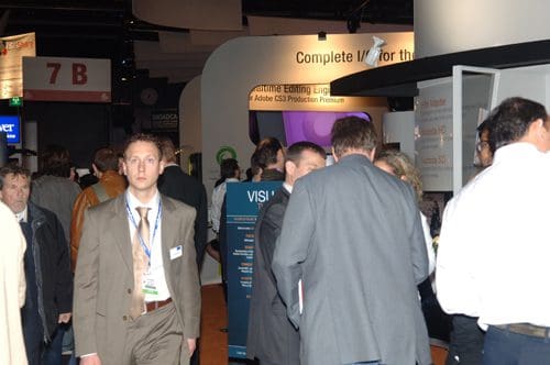 A busy trade show floor with attendees walking between exhibition booths. Some people are conversing, while others are focused on displays in the background. A man dressed in a suit stands prominently in the foreground.
