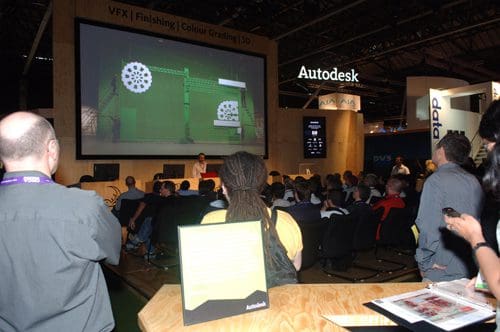 A large audience seated in a conference hall, watching a presentation on a big screen showcasing a digital animation. An information display for Autodesk is visible in the foreground, alongside attendees engaged in discussion.