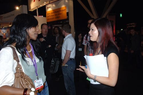 Two women engaged in conversation at a professional event. One woman, with long dark hair and wearing a light outfit, smiles while listening. The other woman, with shoulder-length red hair dressed in black, appears animated, holding a notepad and pen. In the background, attendees are visible, creating a lively atmosphere.