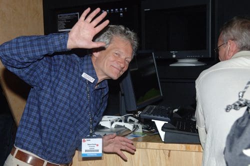 A man with gray hair and a blue checked shirt waves at the camera, standing in front of a computer station. He wears a name tag around his neck, while another person sits at the desk behind him, partially obscured.