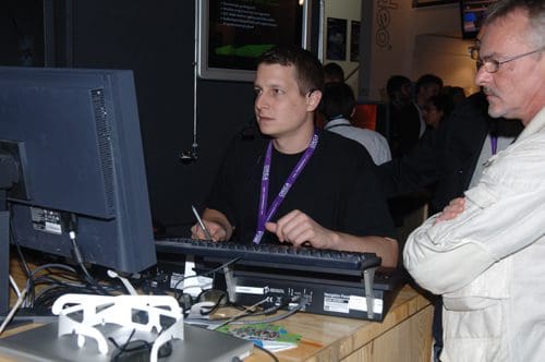 A young man focused on his computer screen while using a keyboard, with a notepad in hand. An older man stands next to him, observing the computer setup at a busy exhibition.