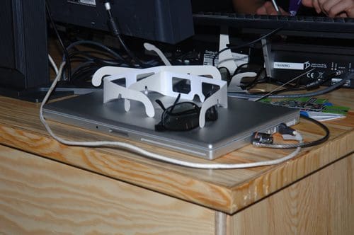 A pair of 3D glasses and a white plastic holder for them resting on a silver laptop. The laptop is placed on a wooden desk, surrounded by cables and a computer monitor in the background.