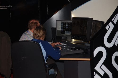 Two individuals collaborating at a computer station, focused on multiple monitors displaying digital content. One person, an adult with light brown hair, is seated behind a child with short hair, both engaged in a tech-related task.