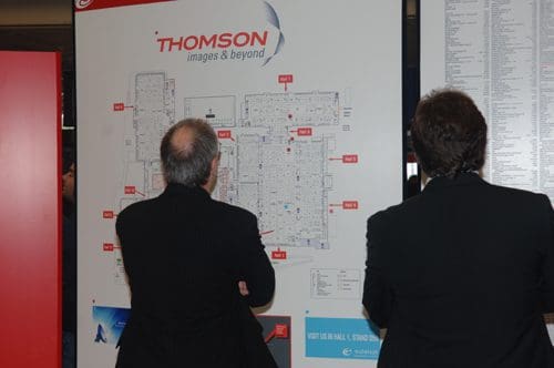 Two men in black suits standing with their backs to the camera, studying a large map on a wall labeled "THOMSON images & beyond." The map shows a detailed layout of a building or venue.