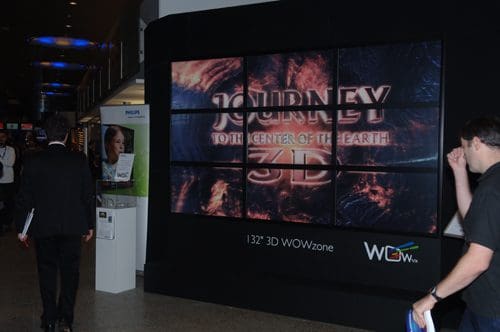 A digital display showcasing the title "Journey to the Center of the Earth 3D" on a large screen in a public space. Two people walk by, one carrying a folder, with promotional signage visible nearby.