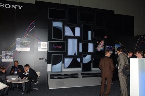 A modern exhibition space featuring a large display wall with multiple screens showcasing digital content. In the foreground, groups of people engage in conversation around tables, surrounded by a sleek, professional atmosphere.