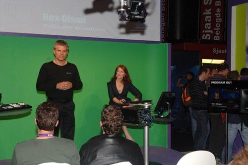 A man in a black shirt stands on stage next to a woman in a black jacket, presenting to an audience. A green screen and equipment are visible in the background, while attendees observe the demonstration.
