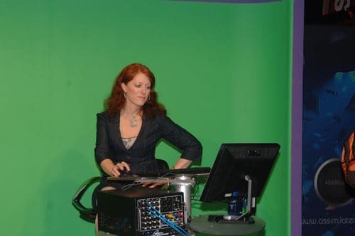 A woman with long red hair wearing a dark blazer stands by a computer setup against a green screen backdrop, engaged in a task. The workspace includes technical equipment and a monitor, showcasing a professional environment.