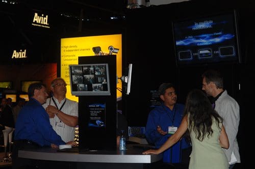 Five individuals engaged in a discussion at a trade show booth with an Avid logo in the background. A monitor displays images, and the atmosphere is casual with a focus on professional networking.