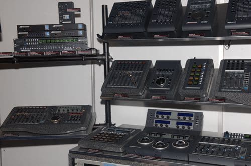 A display of various lighting control panels and equipment on black shelves. The panels feature buttons, knobs, and screens, designed for managing stage lighting. Labels are visible on the shelves for each item.