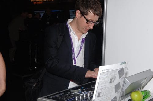A young man in a black blazer and glasses is focused on a DJ mixing console at an event. He has a shoulder bag and a name badge visible around his neck, with an apple and a brochure on the table beside him.