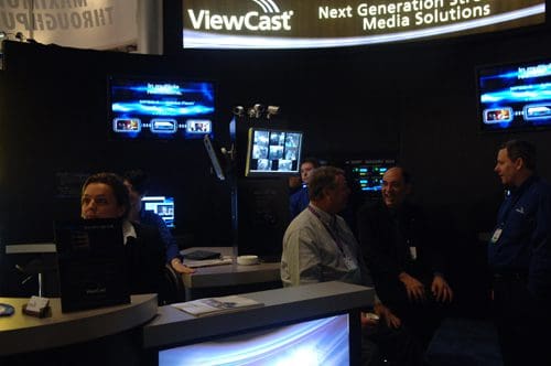 A booth at a trade show featuring ViewCast branding. Individuals are engaged in conversation near display screens showcasing media solutions. The backdrop is dark, enhancing the visibility of the technology and promotional materials.