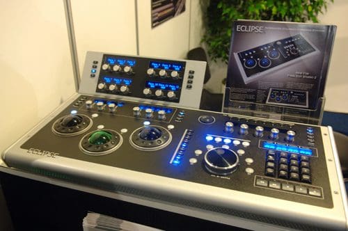 A modern audio mixer with various controls including knobs, dials, and buttons, featuring illuminated displays. It is displayed on a black surface in a well-lit setting, surrounded by a white backdrop and green foliage.