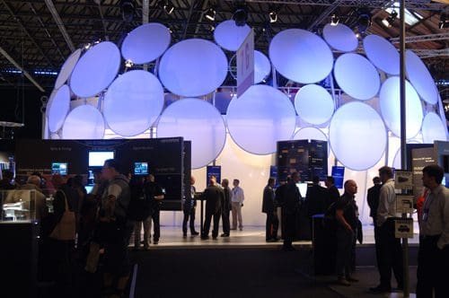 An exhibition booth featuring large white circular installations, illuminated with blue lighting. Attendees, dressed in business attire, engage in discussions and explore displays showcasing technology products around the booth.