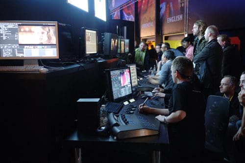 A group of people observing a live editing demo at a technology event, with monitors displaying video editing software. Several technicians are seated at workstations equipped with control panels and multiple screens, focused on their tasks.