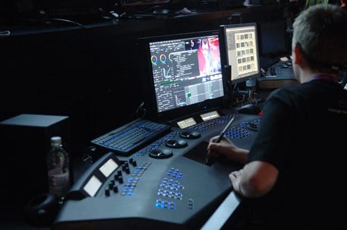 A person working at a control panel with multiple screens displaying graphics and data. The setup includes colorful buttons and knobs, with a water bottle visible nearby in a dimly lit environment.