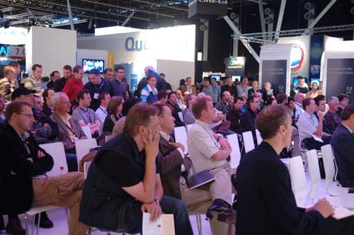A large audience of diverse individuals seated in a convention hall, attentively listening to a speaker at an event. Bright lighting highlights the modern exhibition space with various screens in the background.