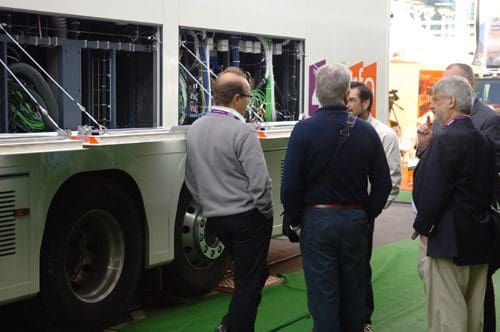 A group of professionals discussing a large vehicle, focusing on its rear components. The vehicle, possibly a bus, features visible mechanical parts and cables. The setting appears to be an exhibition, with people in business attire.